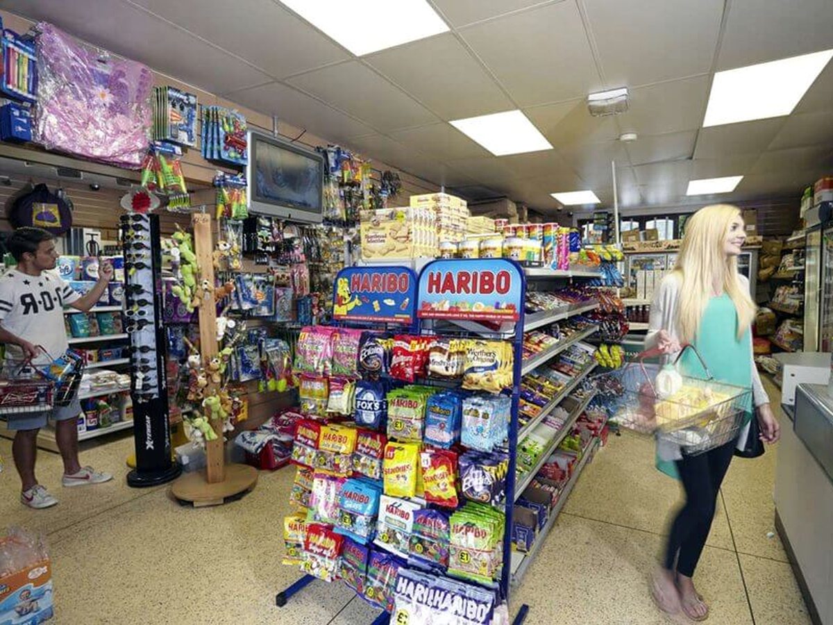 Shoppers with baskets in a small convenience store, beside a Haribo stand and shelves of biscuits, drinks and holiday essentials.