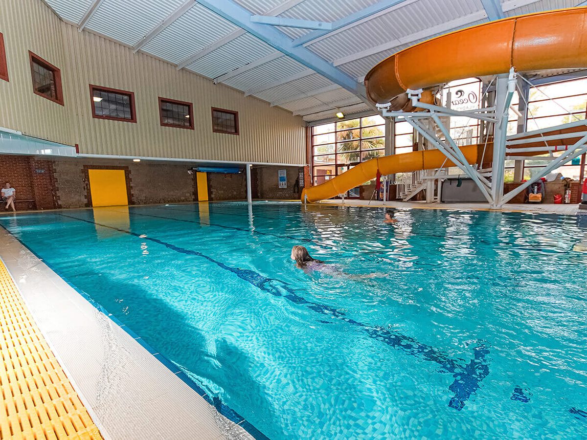 Indoor pool hall with a long blue-tiled pool, a swimmer mid-length, an orange enclosed flume curving down from a steel tower and high windows along one wall.