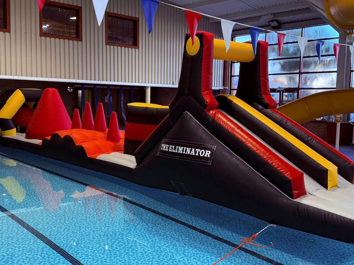 A long black, red and yellow inflatable obstacle course with slides, ramps and climbing sections floating in the indoor pool, with bunting strung overhead.