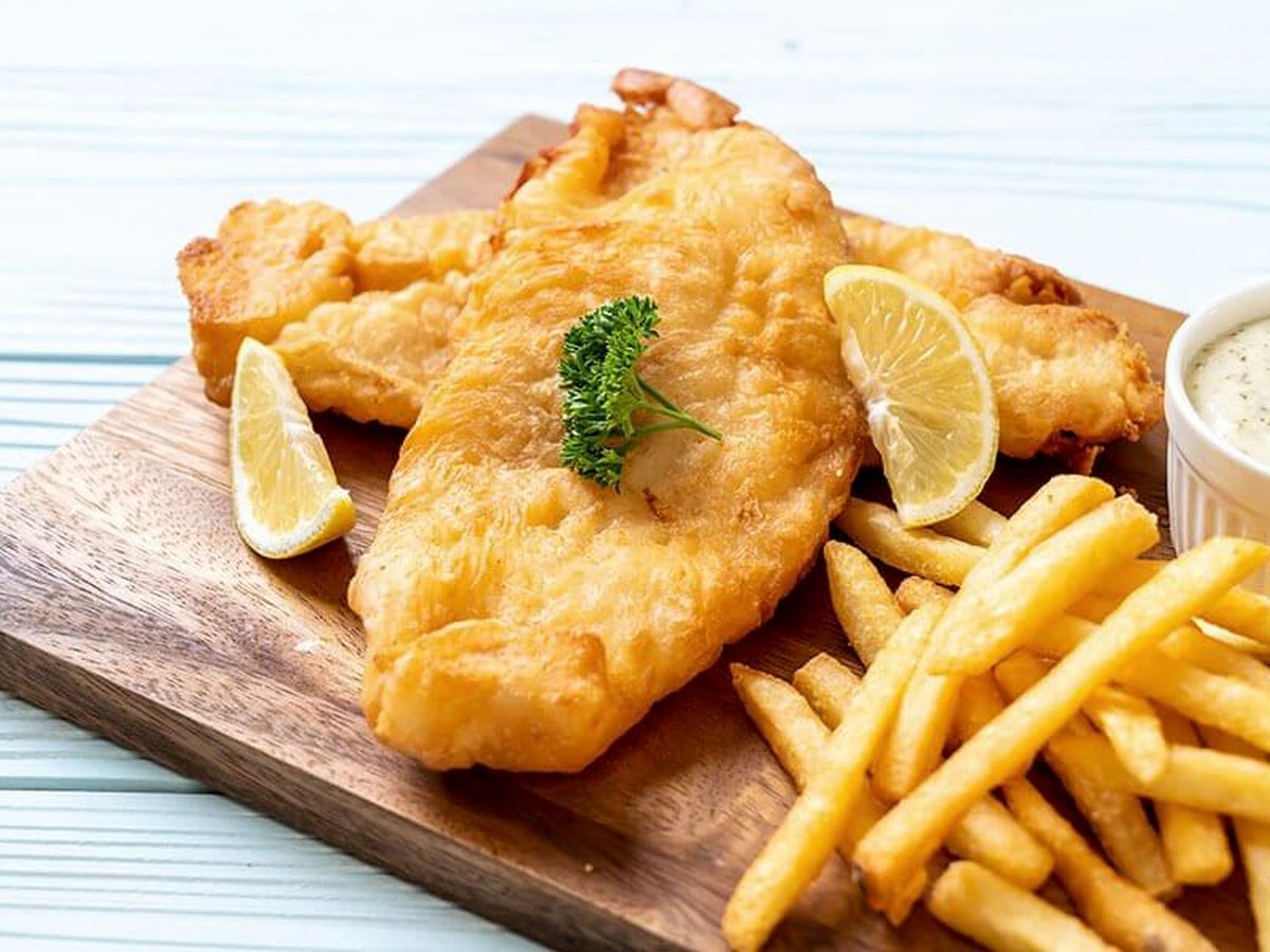 A wooden board holding a piece of battered fish, a pile of chips, lemon wedges and a pot of tartare sauce.