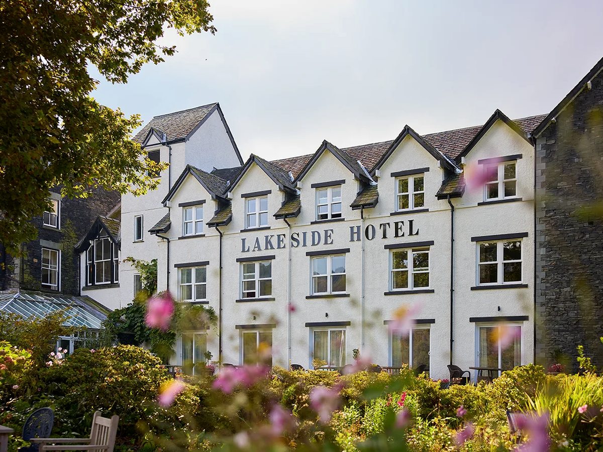 Three-storey white rendered hotel with gabled dormer windows and Lakeside Hotel lettering across the wall, behind flowering shrubs and a garden bench.