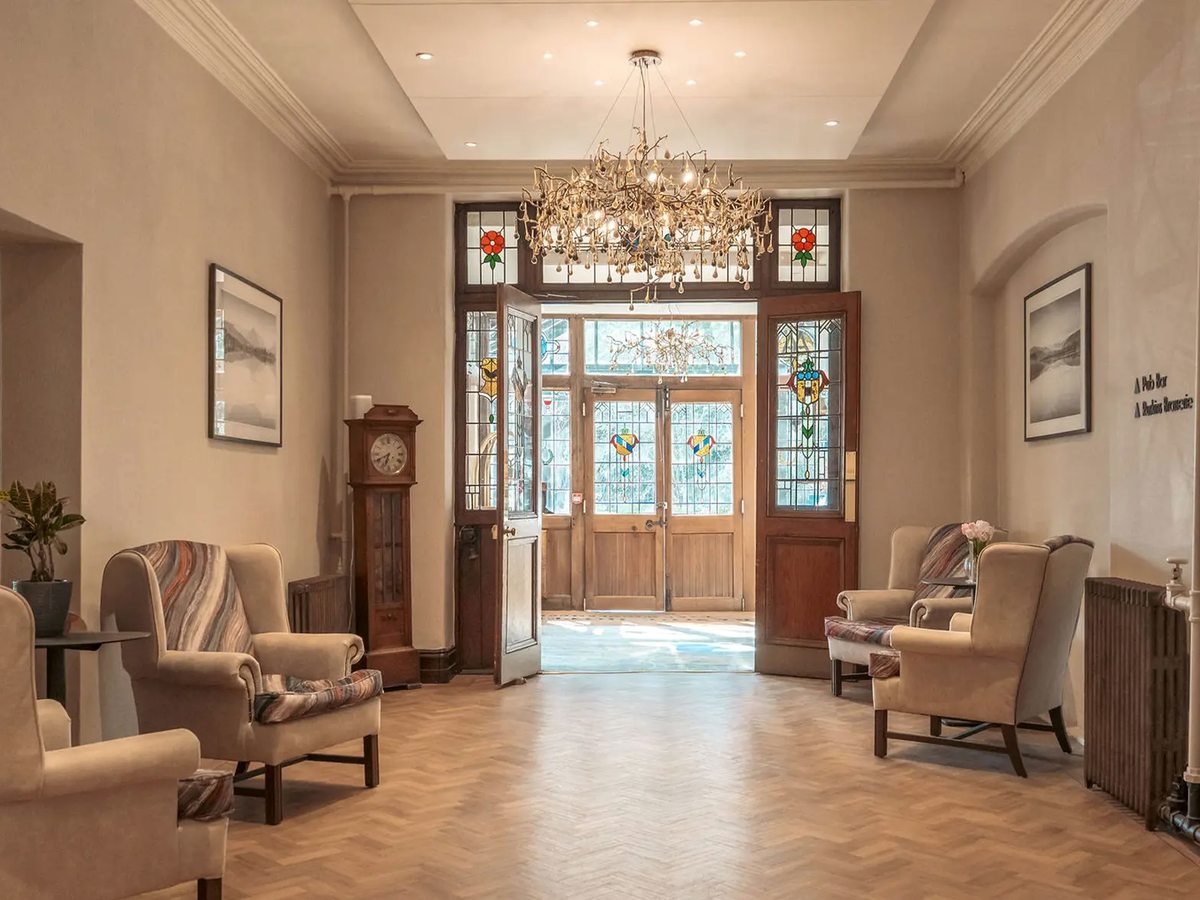 Entrance hall with parquet flooring, a branch-shaped chandelier, a grandfather clock, wing-back armchairs and stained-glass doors standing open.