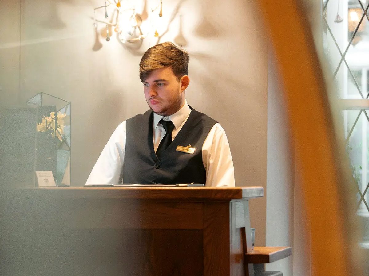 A member of staff in a waistcoat and tie behind a polished wooden reception desk, under a chandelier beside leaded windows.