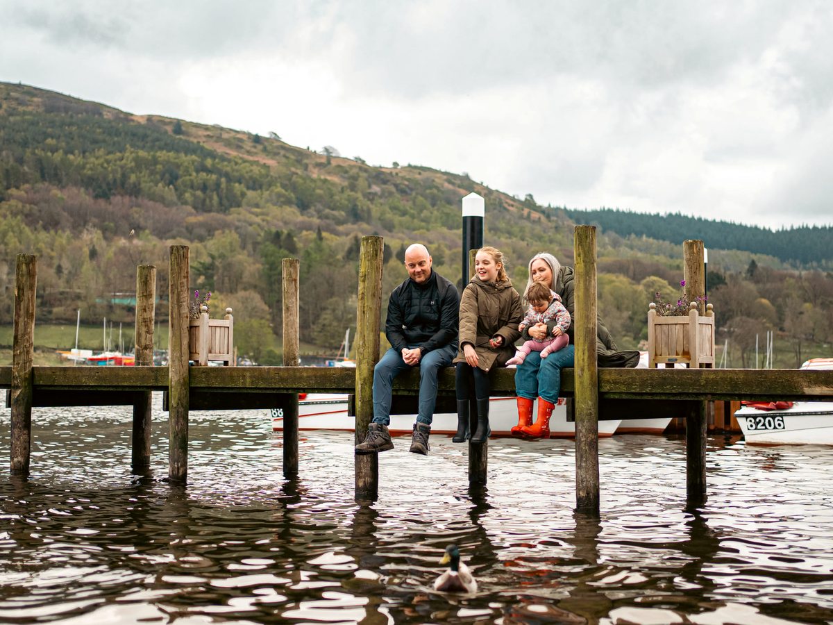 Two adults, a girl and a toddler sitting along the edge of a wooden jetty above the water, a duck swimming below and a wooded fell behind.