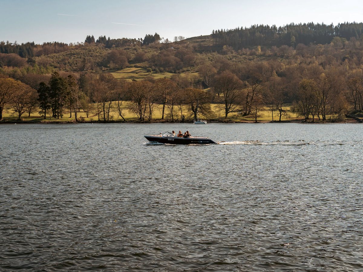 A small motorboat crossing open water, with a wooded shoreline and low green hills behind.