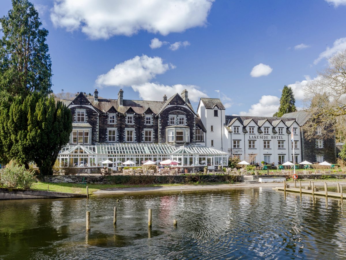 The hotel seen across the water on a sunny day, a long glass conservatory and parasol-shaded terrace along its front, slate and white buildings behind and mooring posts in the shallows.
