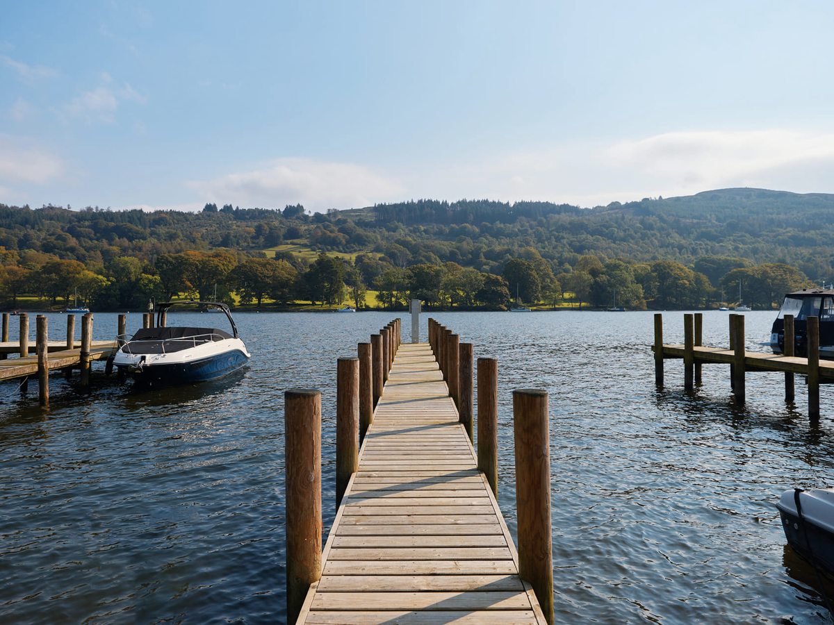 A wooden jetty running out into the lake between mooring posts, motorboats tied up either side and low wooded hills across the water.