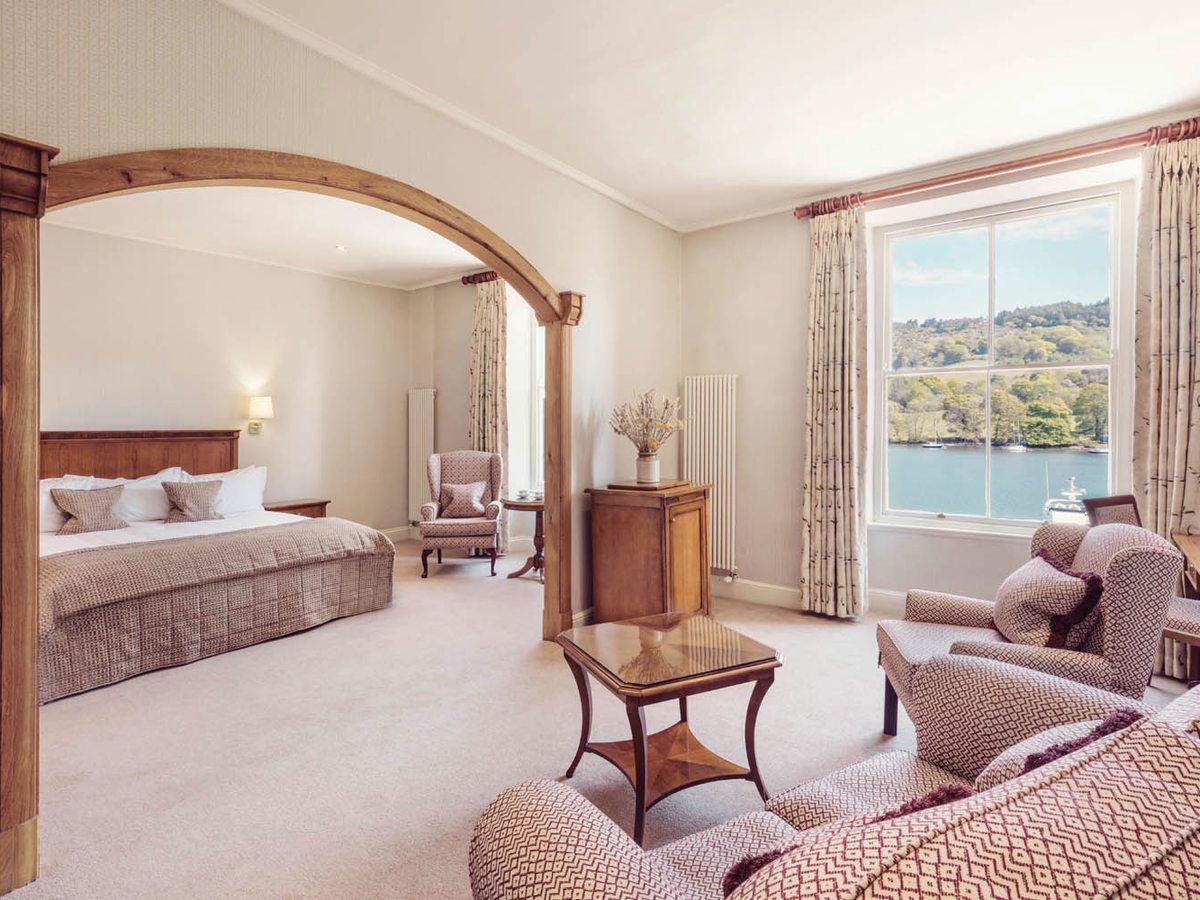 Large room with a double bed set through a wide oak arch, patterned armchairs and a low table by a tall sash window with the lake and its wooded shore beyond.