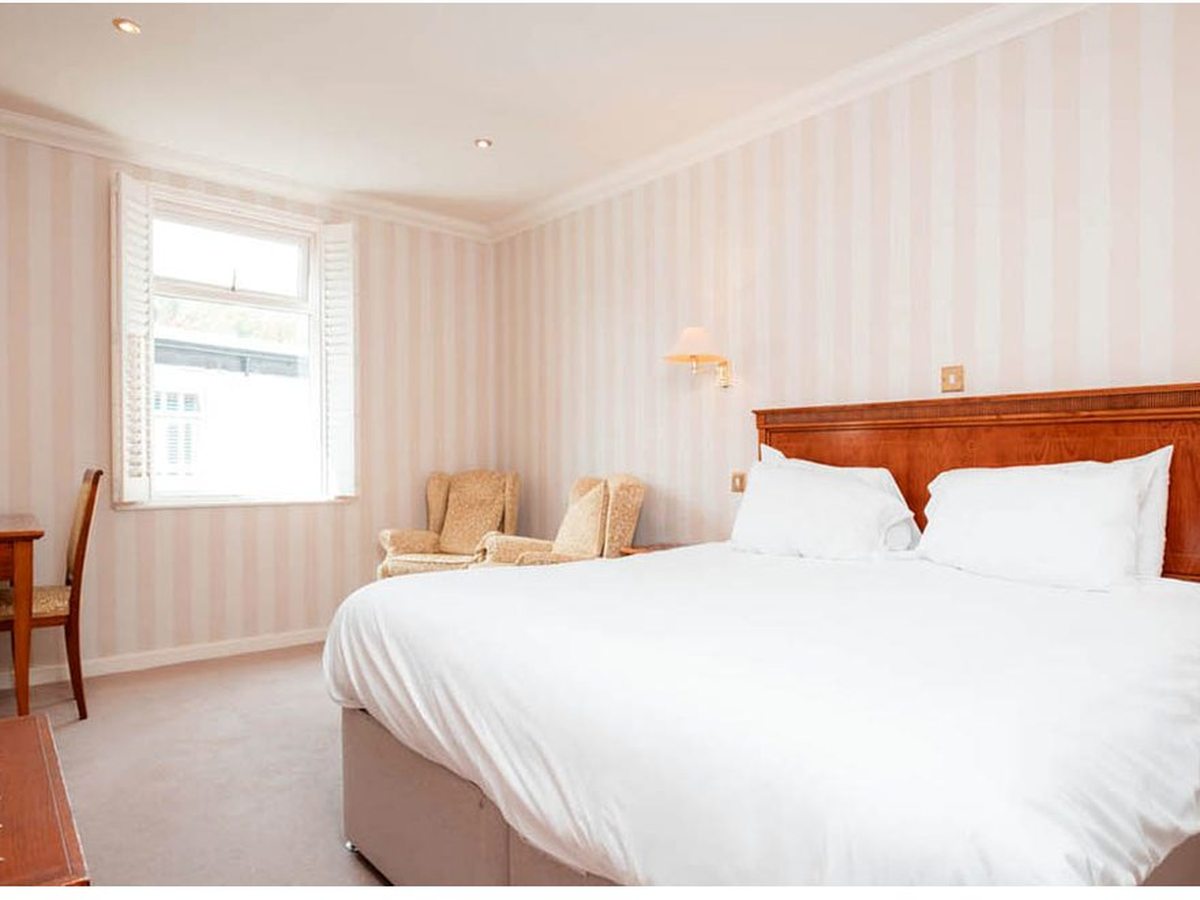 Simple double room with white bedding, a wooden headboard, striped wallpaper, two small armchairs and a writing desk by a shuttered window.