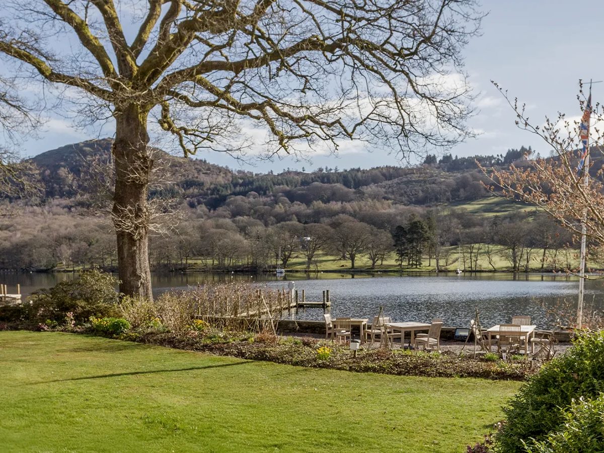 Mown lawn and planted borders under a large bare oak, with wooden tables at the shore, a jetty and the lake backed by wooded hills.