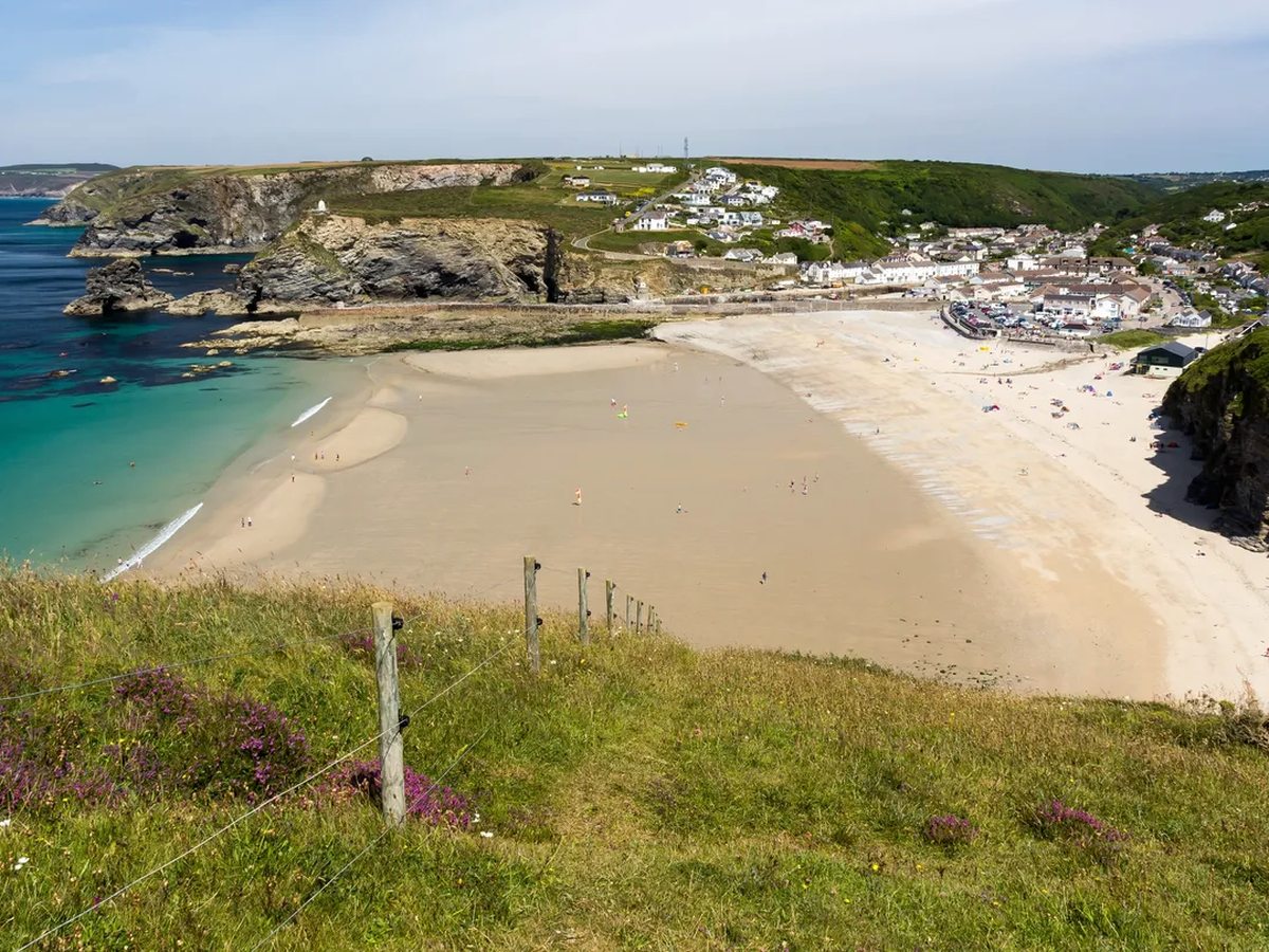 A wide sandy beach in a cove seen from a grassy clifftop, with turquoise water, dark cliffs to one side and a village of white houses behind.