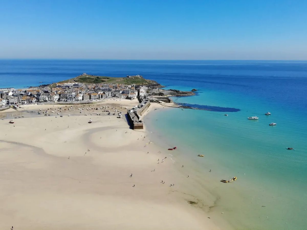 Aerial view of a sandy beach and a harbour town on a headland, with clear turquoise sea and small boats offshore.