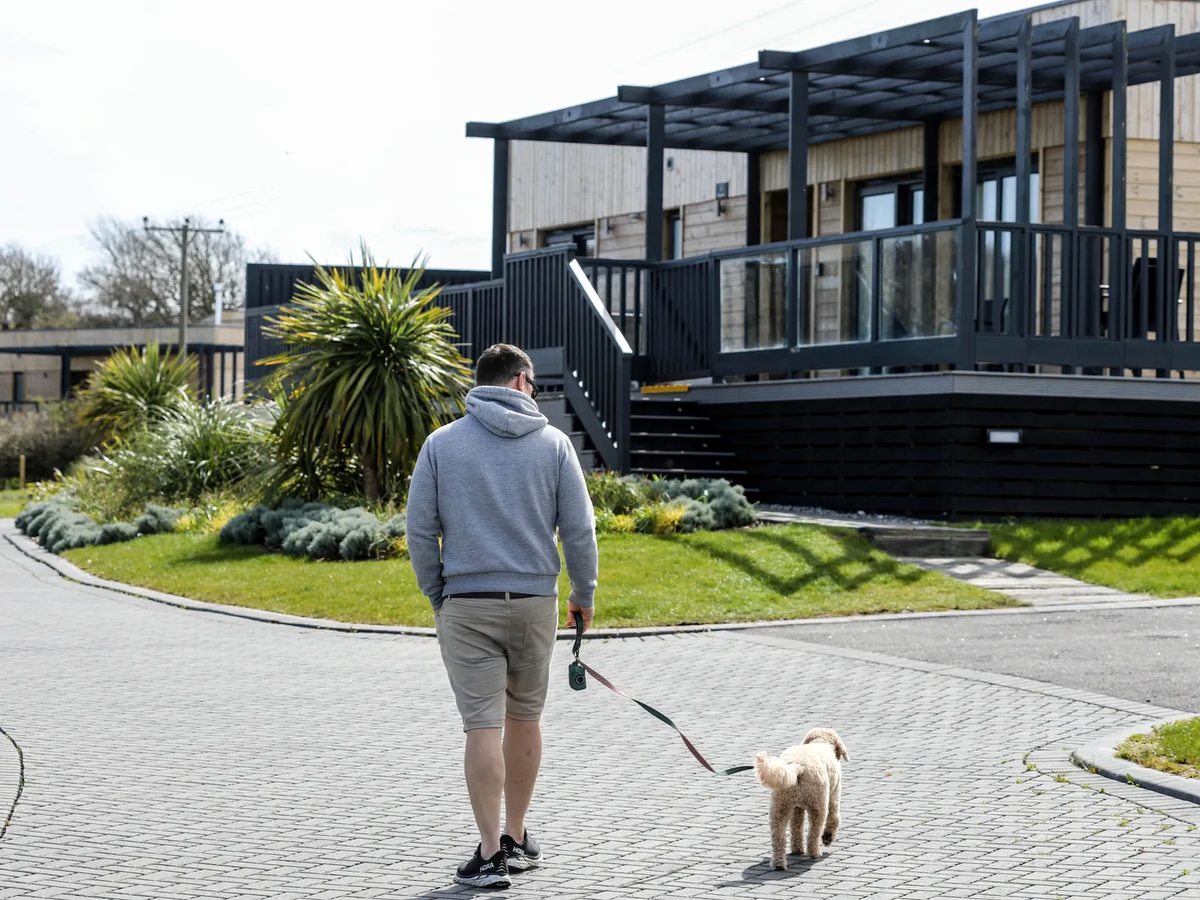 A man in shorts walks a small white dog along a paved lane between timber lodges with planted borders and lawns.