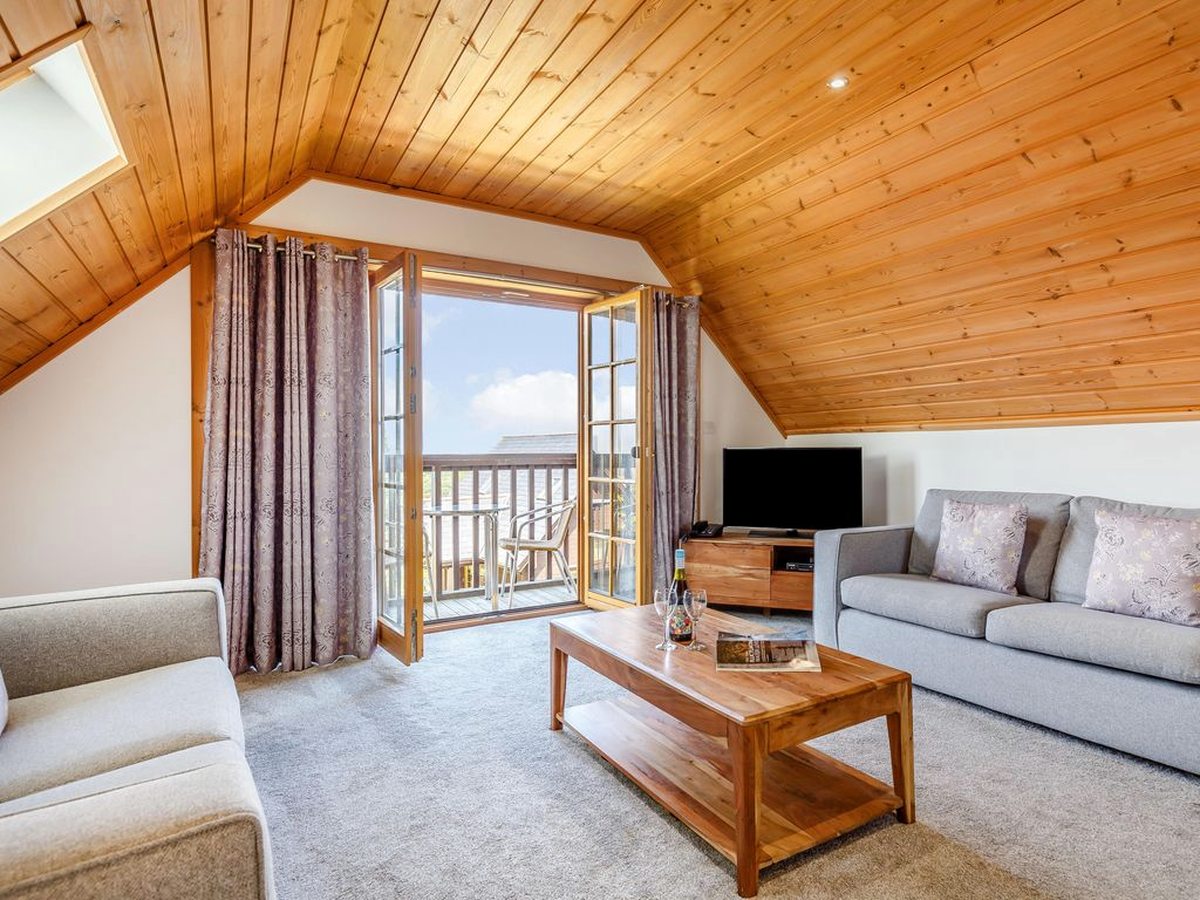 A lodge living room with a vaulted pine ceiling, pale sofas, a low wooden coffee table and glazed doors onto a balcony.