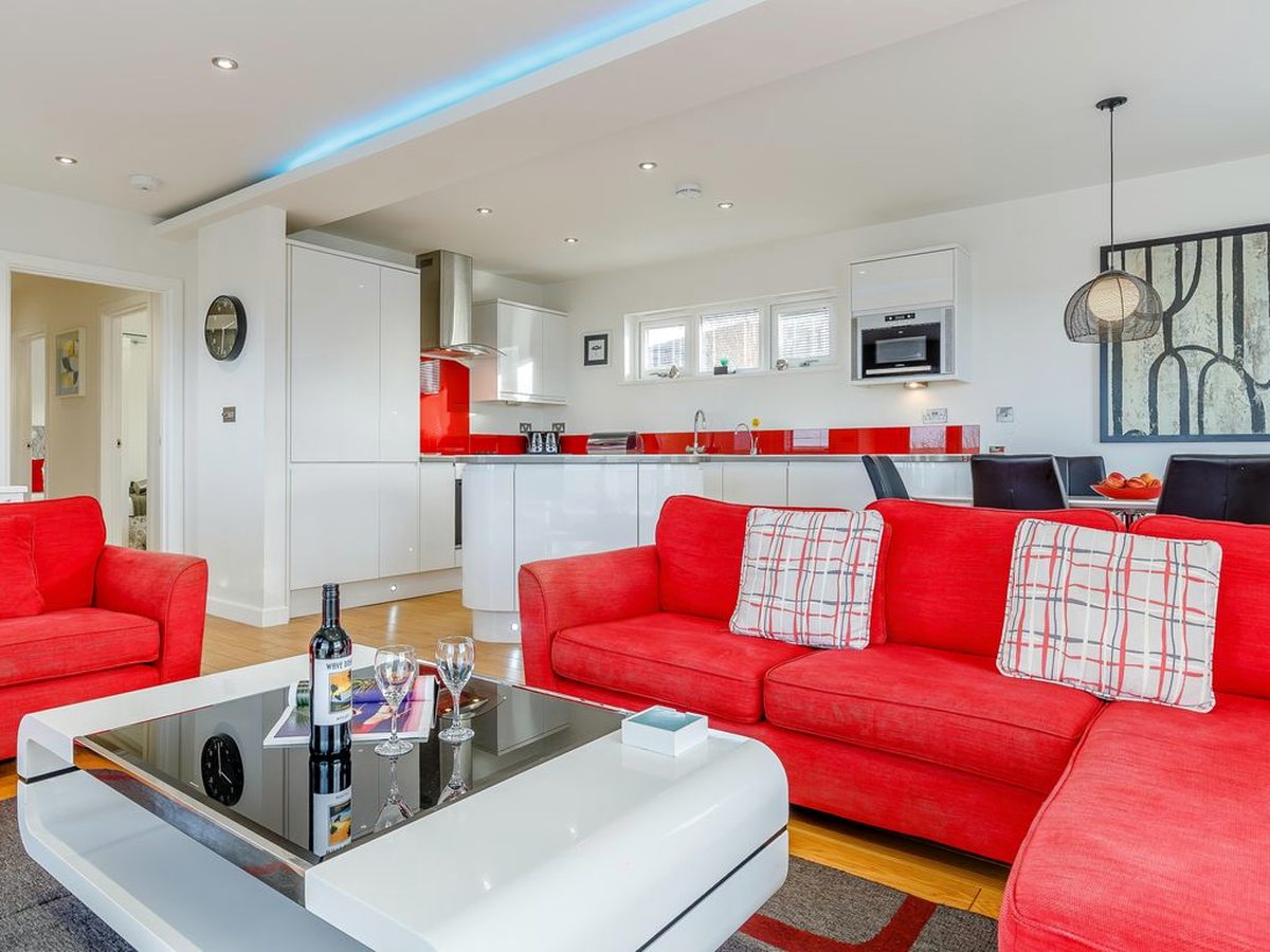 An open-plan lodge interior with red sofas, a white coffee table and a white kitchen with red splashback along the back wall.