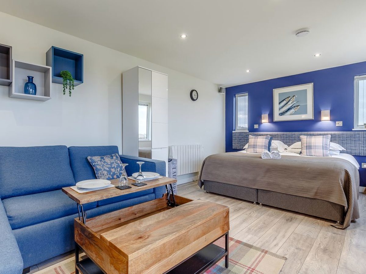 A studio interior with a double bed against a deep blue wall and a blue sofa and wooden coffee table in front of it.
