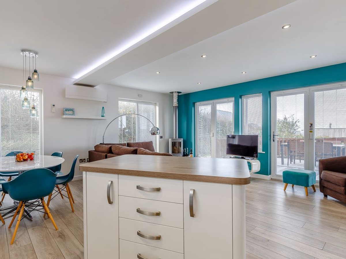 An open-plan kitchen and dining area with white units, a teal splashback, a dining table with turquoise chairs and wide glazed doors.
