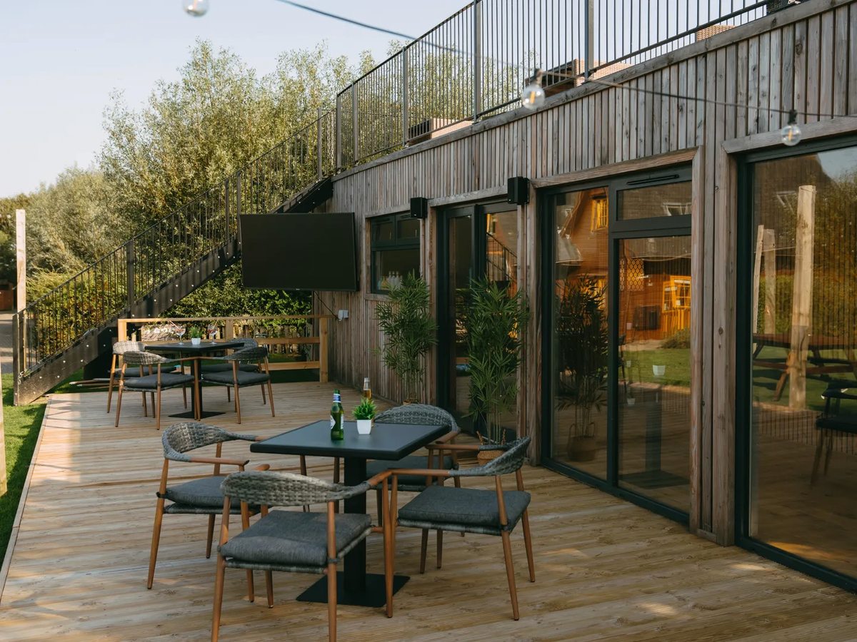 Rattan chairs and dark square tables on a timber deck outside a timber-clad building with sliding glass doors and a balcony above.