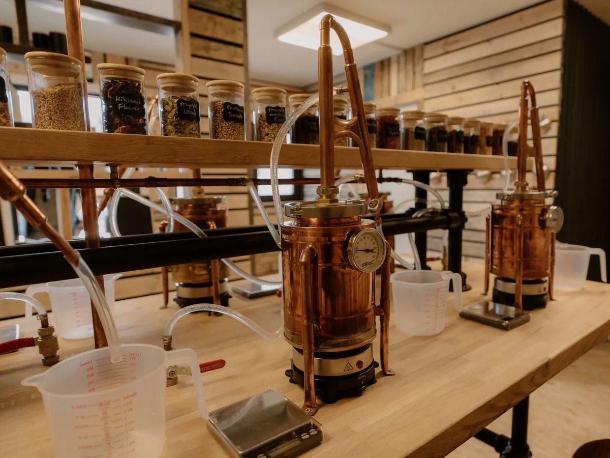 Two small copper gin stills on a wooden bench with measuring jugs beside them and shelves of labelled jars behind.