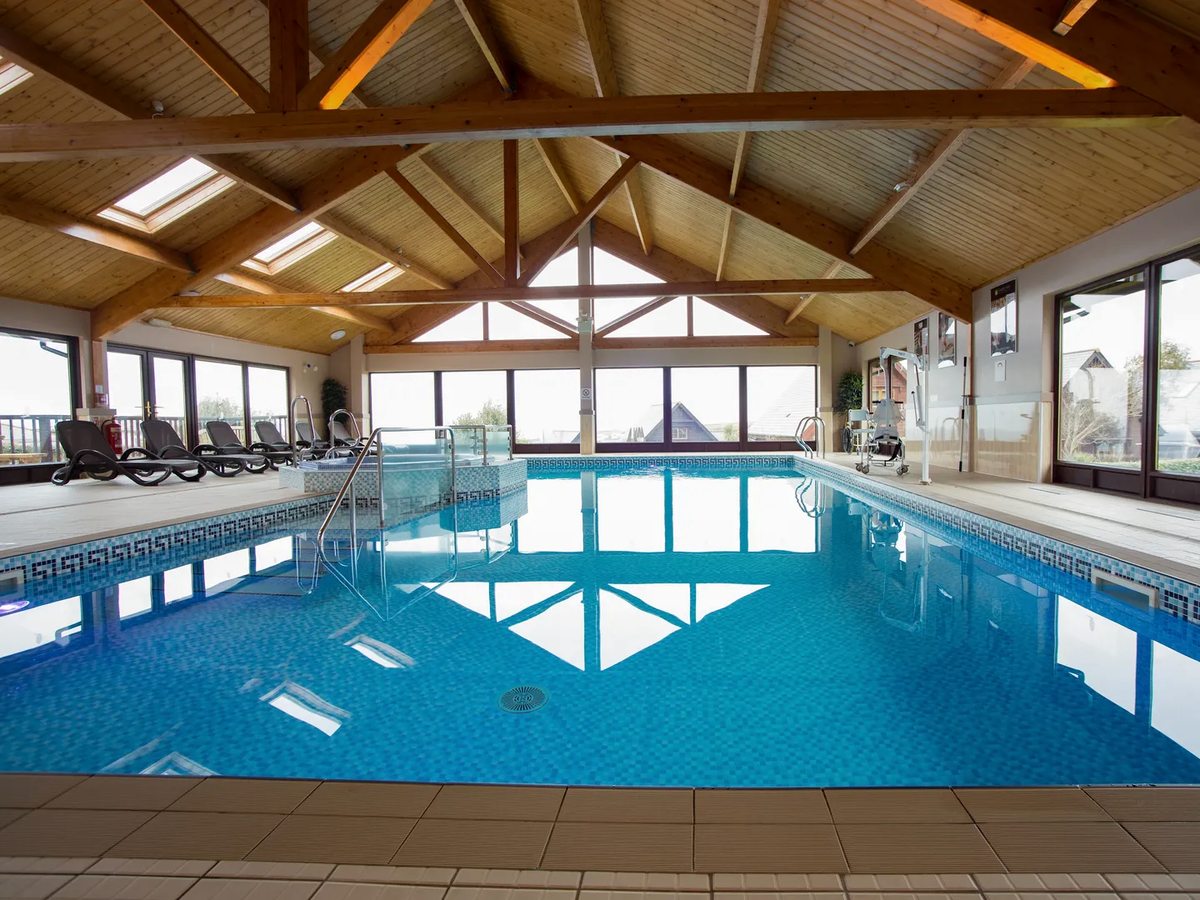 An indoor pool under a pitched timber roof, with blue water, a tiled surround, loungers along one side and tall windows at the far end.