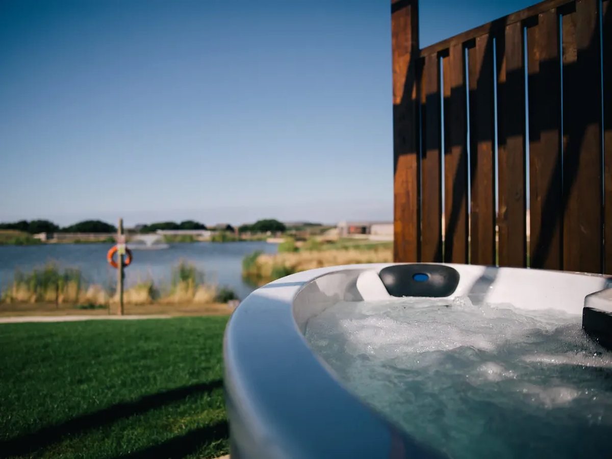 A round hot tub with a moulded rim on a lawn, with a lake, grass banks and a fountain beyond.