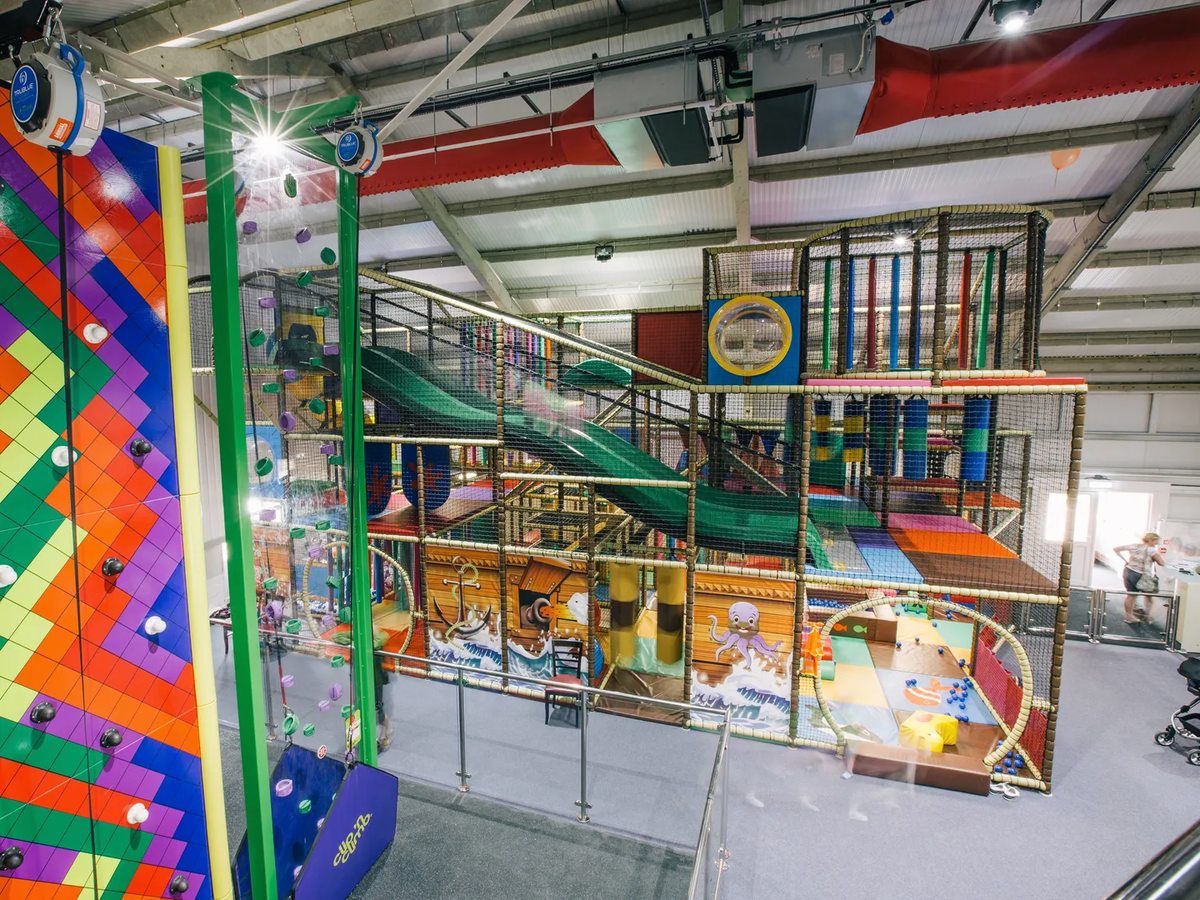 A large multi-level indoor play frame with slides, netting and brightly painted panels inside a high-roofed hall.