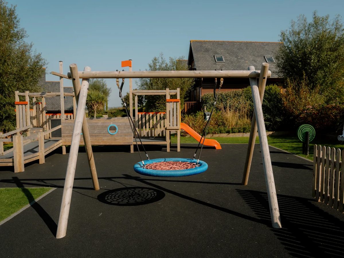 A wooden play frame with swings, a slide and a round basket swing on a black rubber surface, with lodges behind.