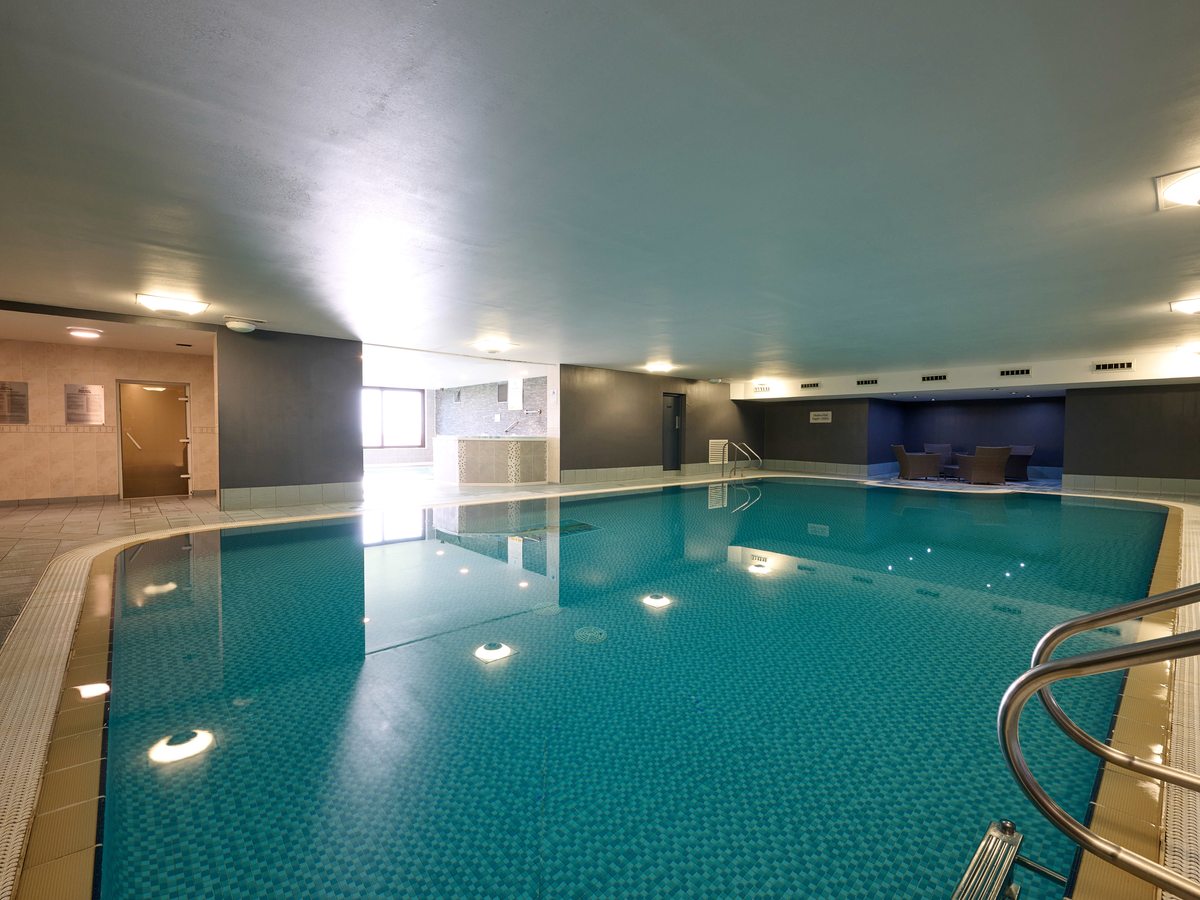 A rectangular indoor swimming pool with underwater lights, a tiled surround, handrails at the corner steps and armchairs at the far end.