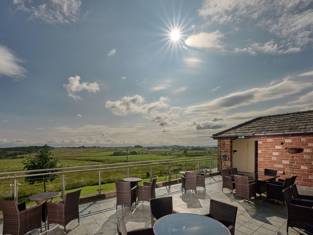 Rattan chairs and tables on a paved terrace behind a glass and steel balustrade, with fields and low hills under a bright sky.