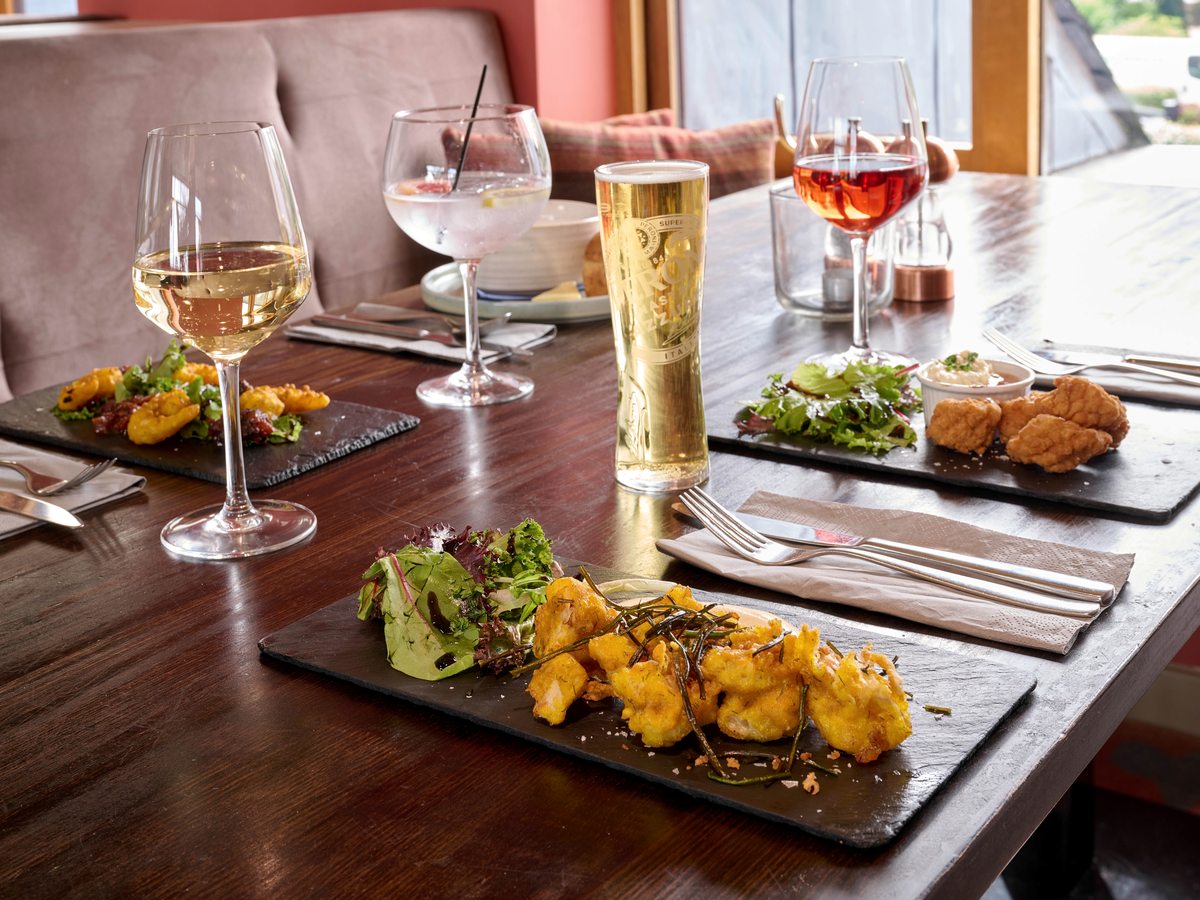 Three slate platters of battered food with salad on a dark wooden table, with a beer, a glass of white wine and two other drinks alongside.