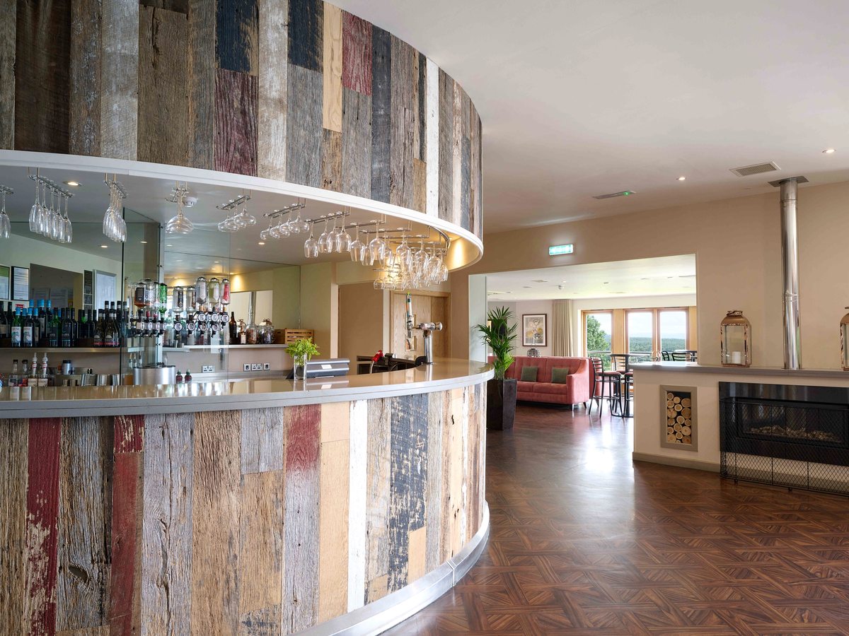 A curved bar counter clad in reclaimed painted timber, glasses hanging above it and a lounge with a wood-burning stove through the opening beyond.