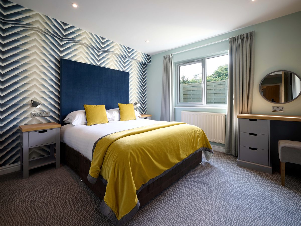 A double bed with a navy padded headboard, yellow throw and cushions against a blue and cream chevron feature wall, with a dressing table by the window.