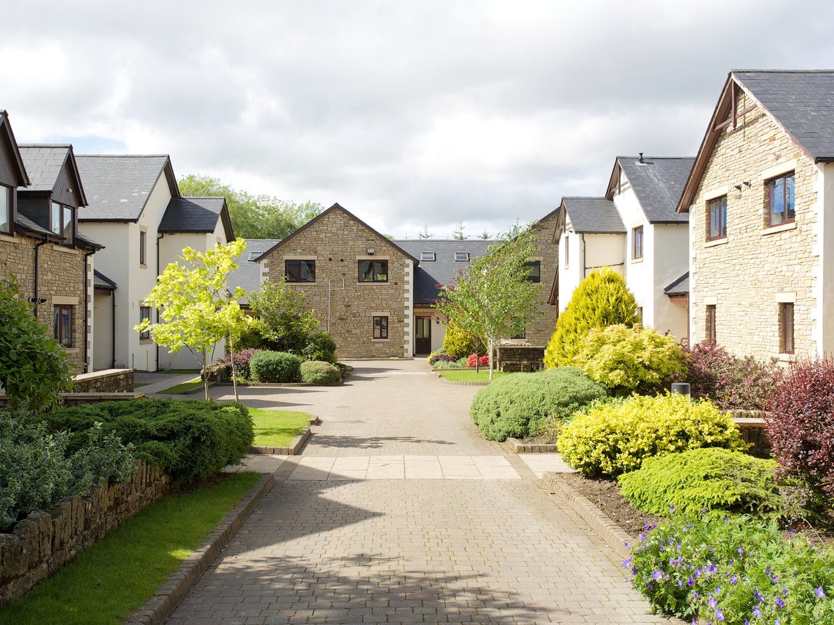 A paved drive running between two-storey stone and rendered holiday homes, with clipped shrubs, small trees and flower beds on either side.