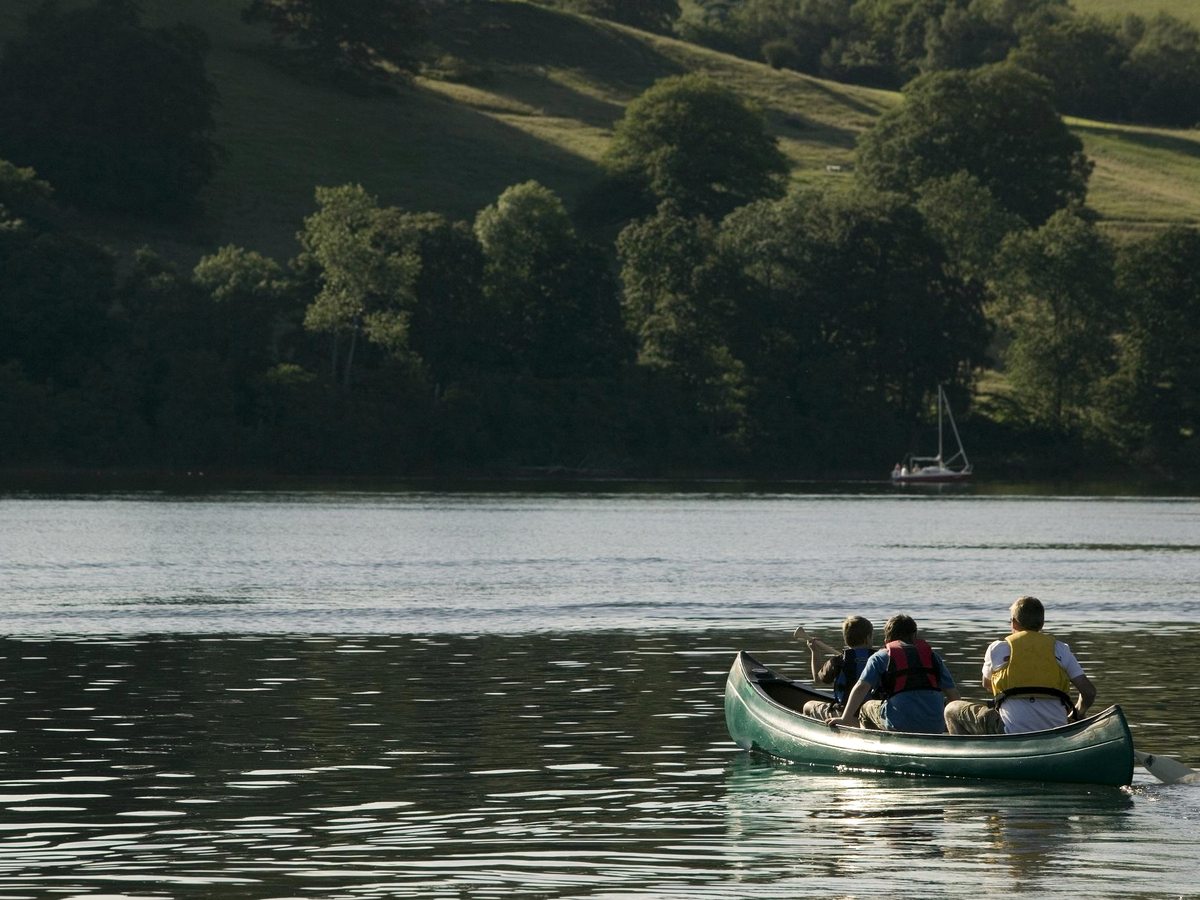 Three people paddling an open green canoe across calm water, with a wooded hillside and a moored yacht behind them.