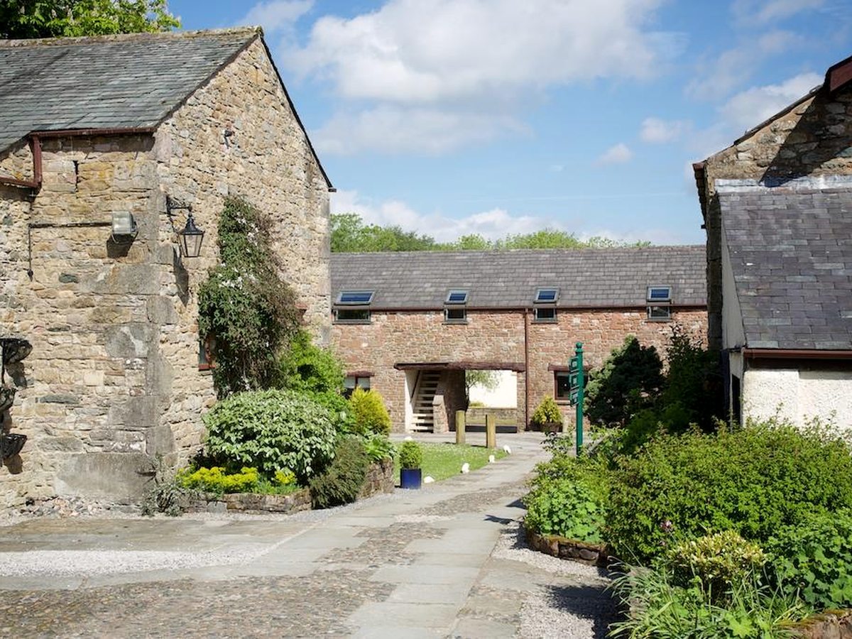 Stone barn buildings with slate roofs around a flagged courtyard, with shrubs, a lantern and a path running between them under a blue sky.