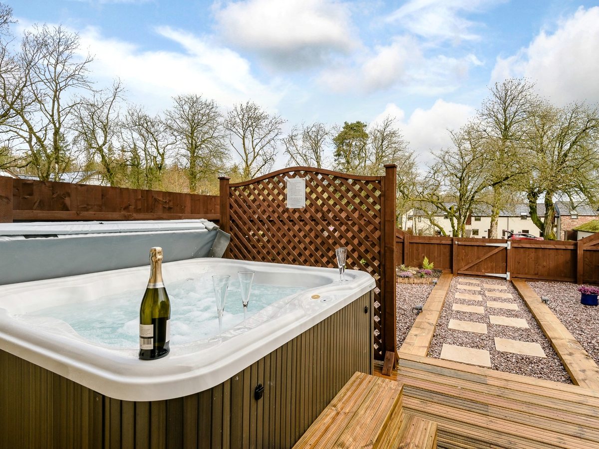 A square hot tub bubbling on a decked terrace enclosed by wooden fencing and trellis, with a paved path and gravel garden alongside.