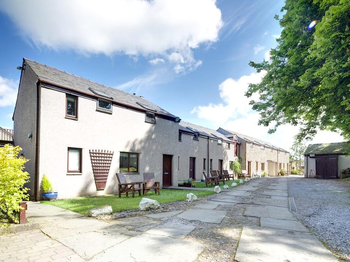 A terrace of rendered stone cottages with front doors opening onto a flagged path, wooden tables and benches outside each one, in sunshine.