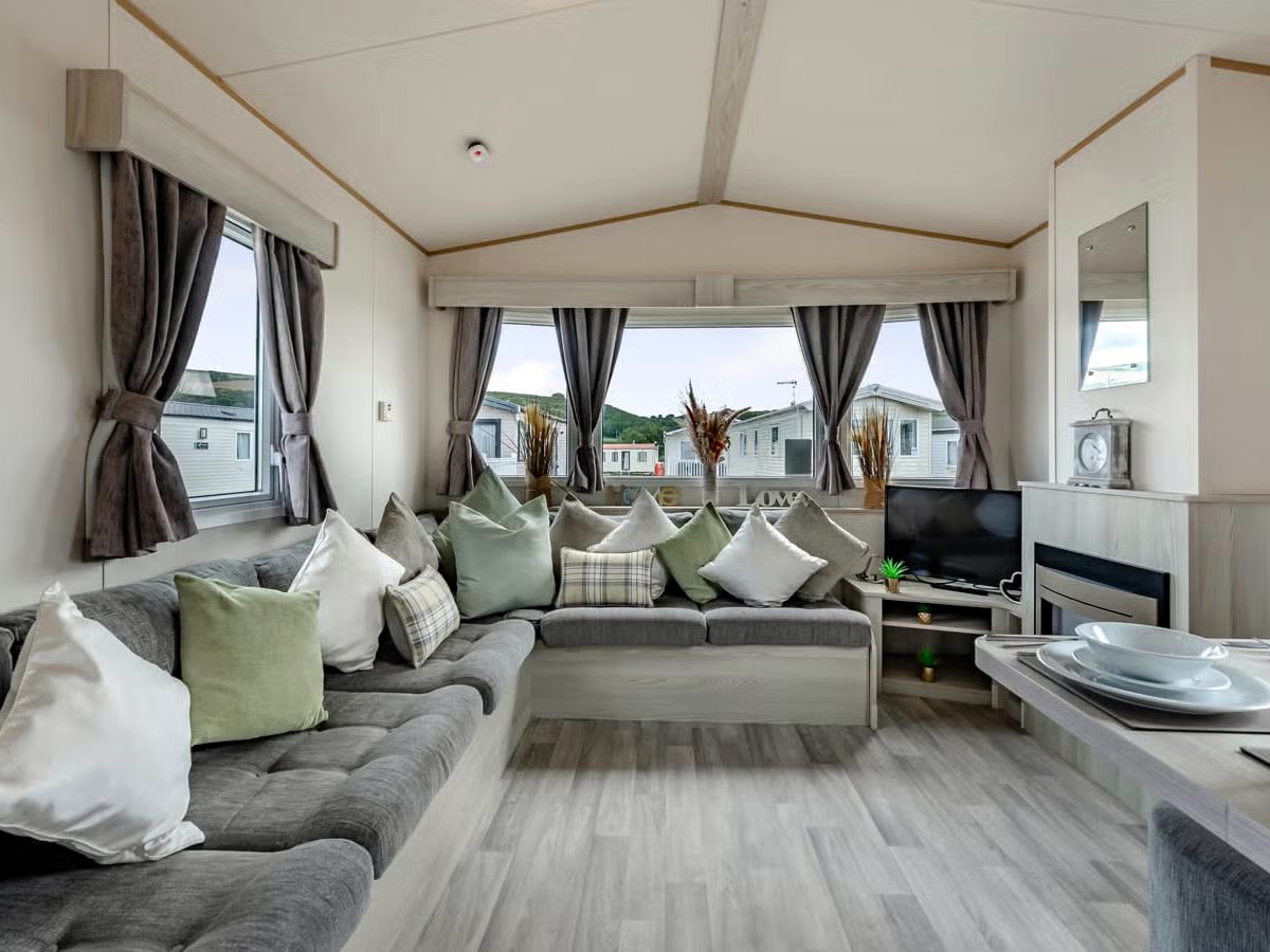 A caravan lounge with a grey corner sofa, cushions, a television and windows onto the pitches outside.