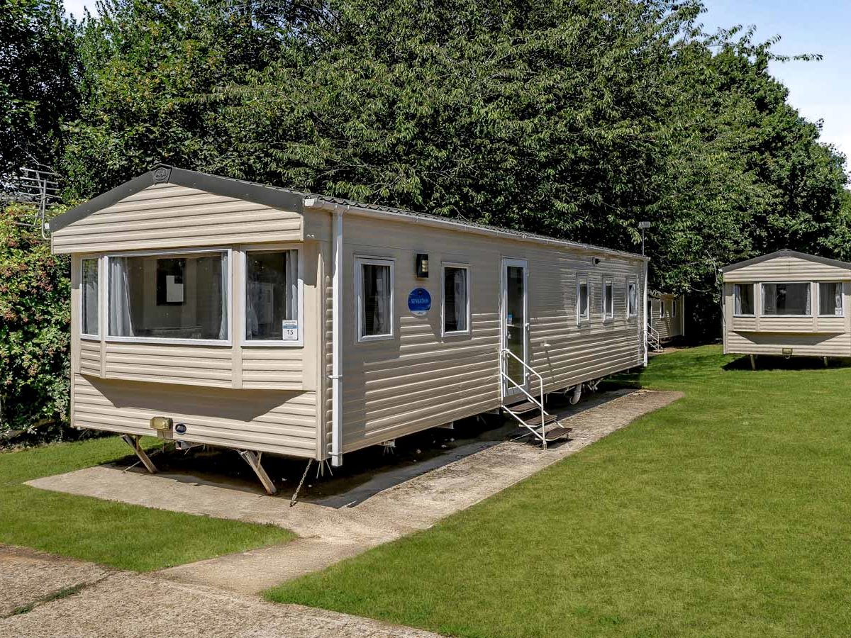 Cream static caravans on concrete pitches in a row, with mown grass and trees behind.