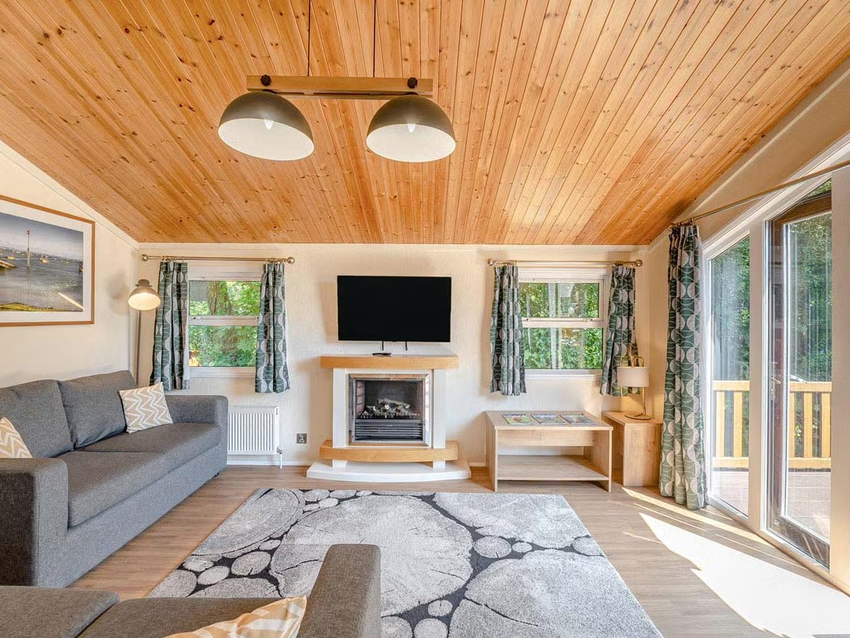 A lodge lounge with a wood-panelled ceiling, grey sofas, a fireplace and glazed doors onto the deck.