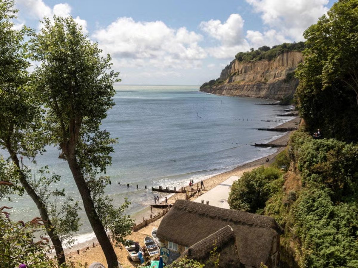 A sandy beach curving below ochre cliffs, with a thatched building at the shore and calm sea.