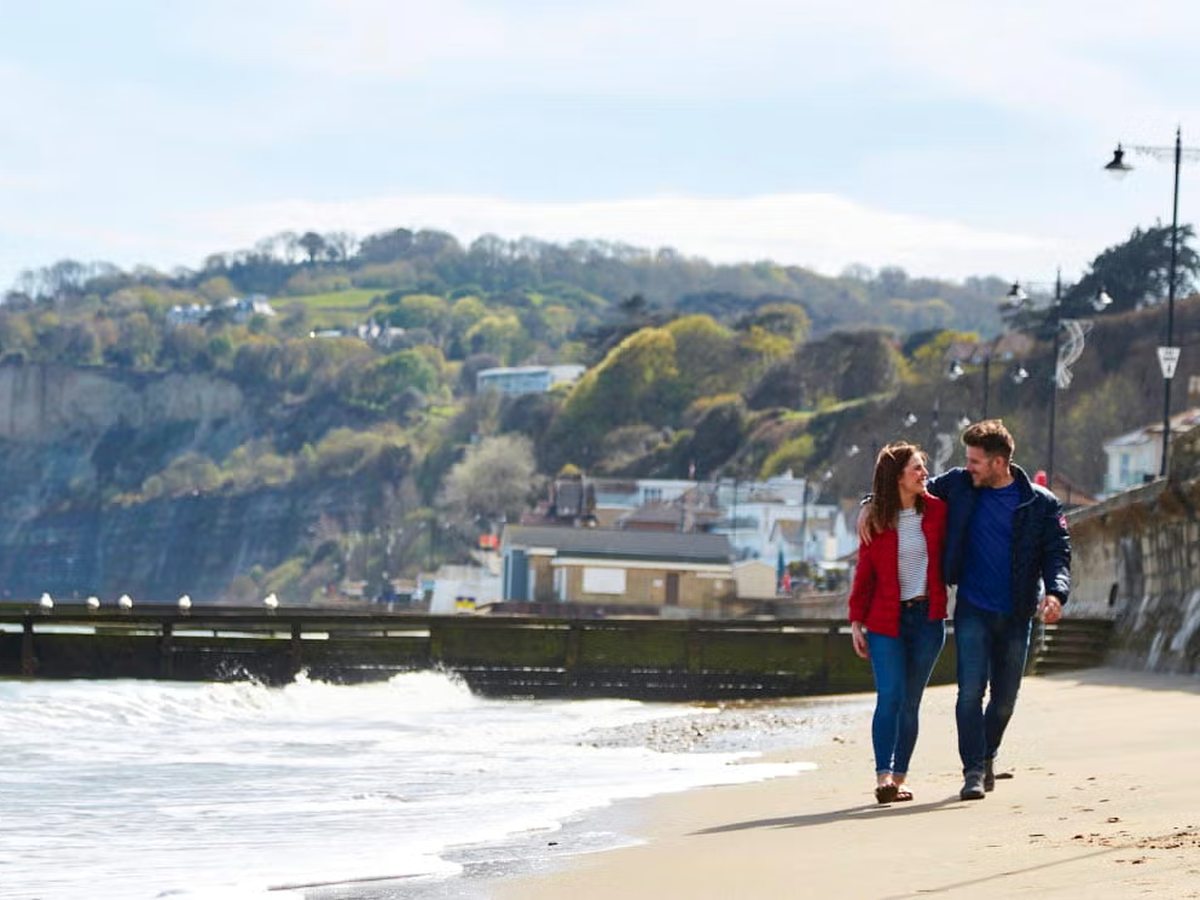 A couple walking along the sand beside a seawall, with seafront buildings and wooded cliffs behind.
