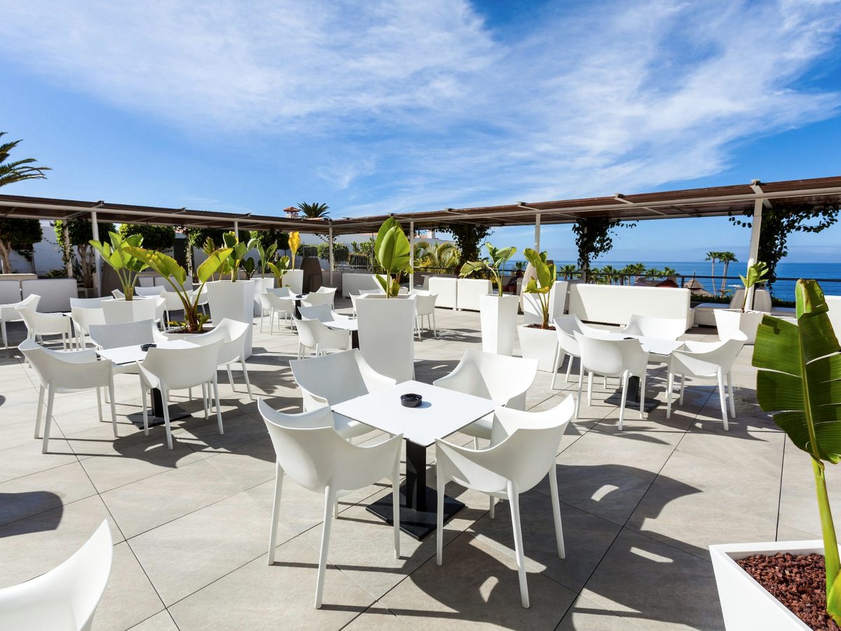 Large open terrace with white tables and chairs, planters of banana palms and a pergola along the far edge above the sea.