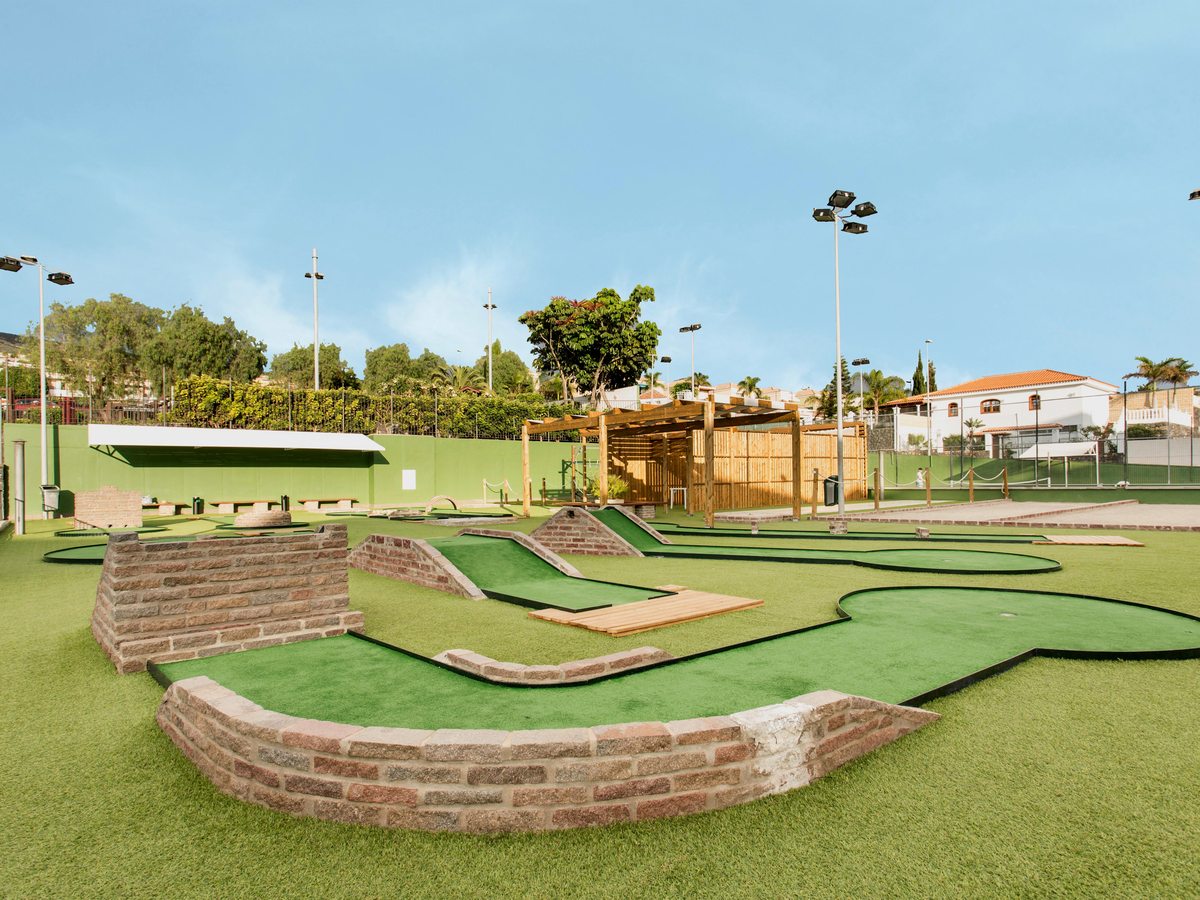 Mini-golf course with artificial turf lanes, brick-edged holes and low wooden obstacles, with floodlights and trees behind.
