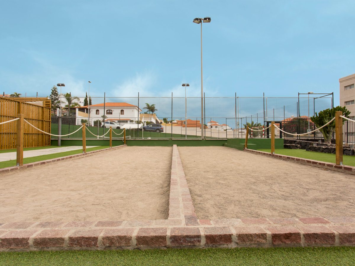 Two sand petanca lanes edged in brick and bounded by rope and wooden posts, with a fenced sports court and houses behind.