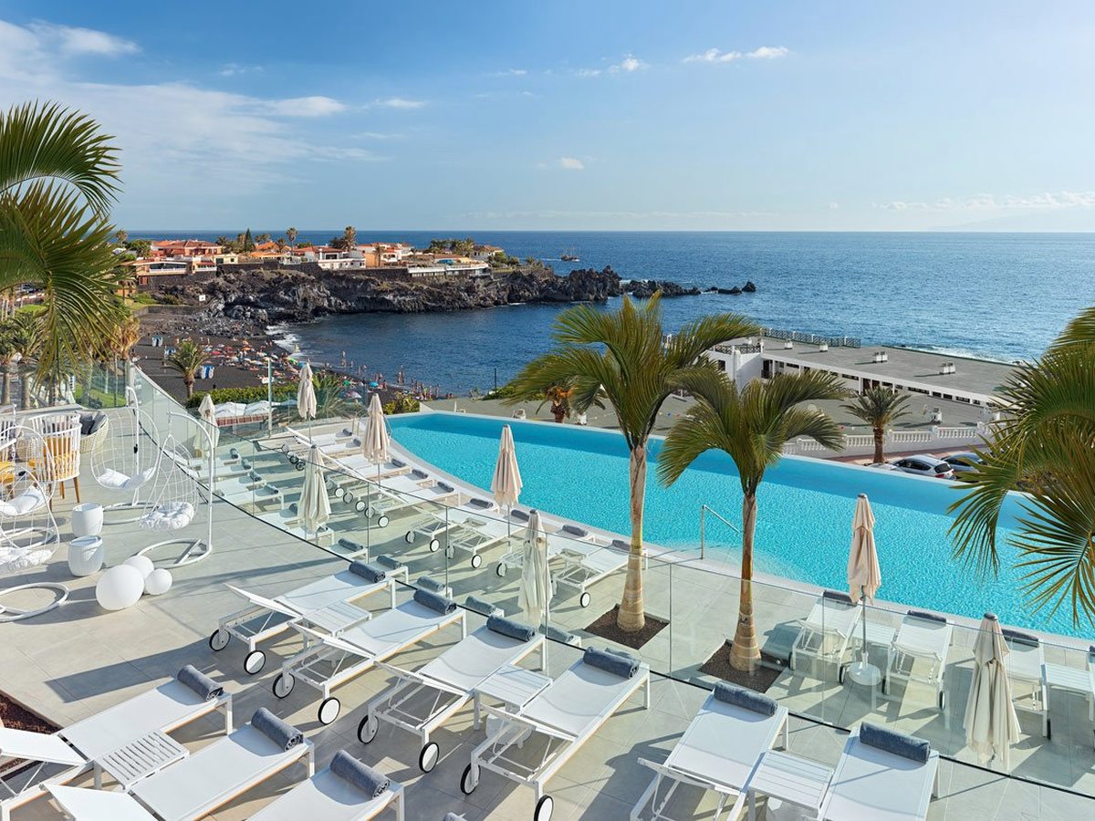Long rectangular infinity pool on a white sun deck lined with loungers and folded parasols, looking over palms to a dark-sand cove and the Atlantic.