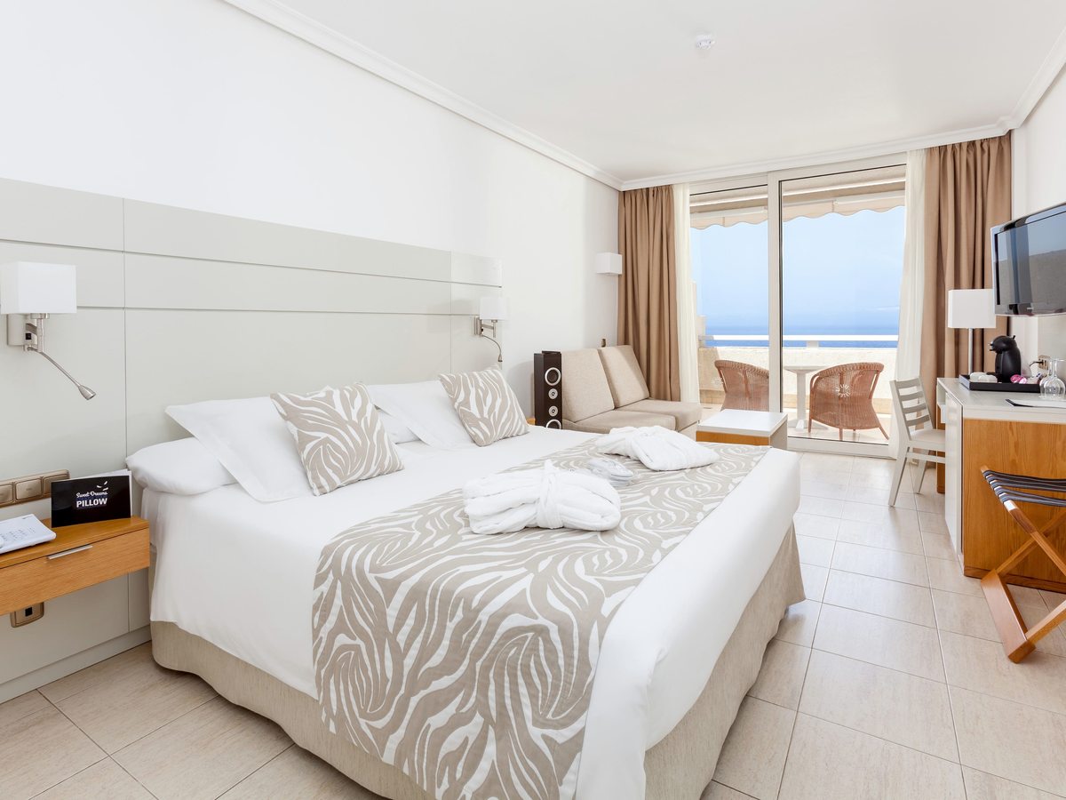 Guest room with a large double bed, a sofa, a desk and sliding doors onto a private terrace with wicker chairs and the sea beyond.