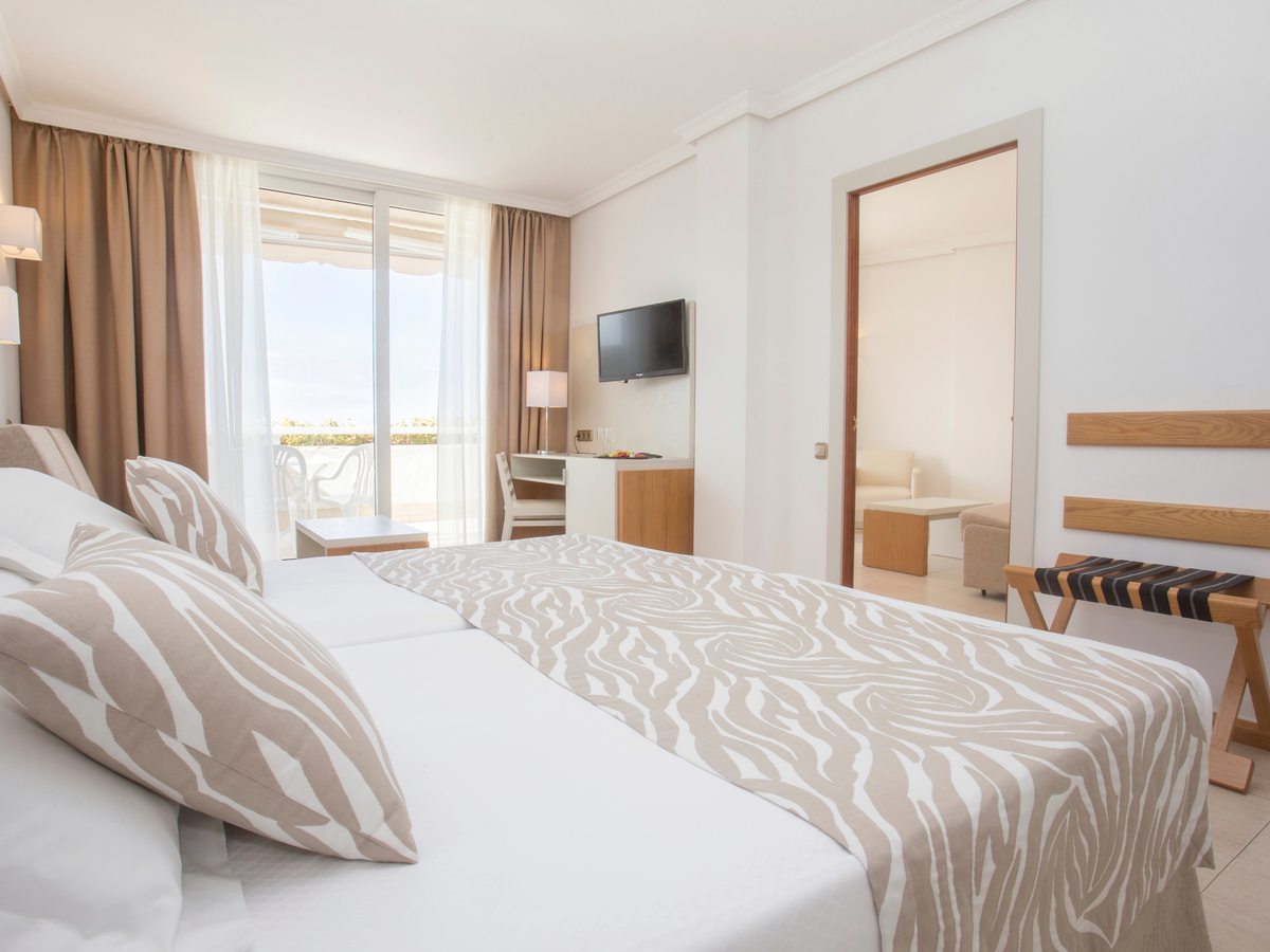 Guest room with a large bed and a wall-mounted television, and a wide doorway through to a seating area with an armchair and a low table.