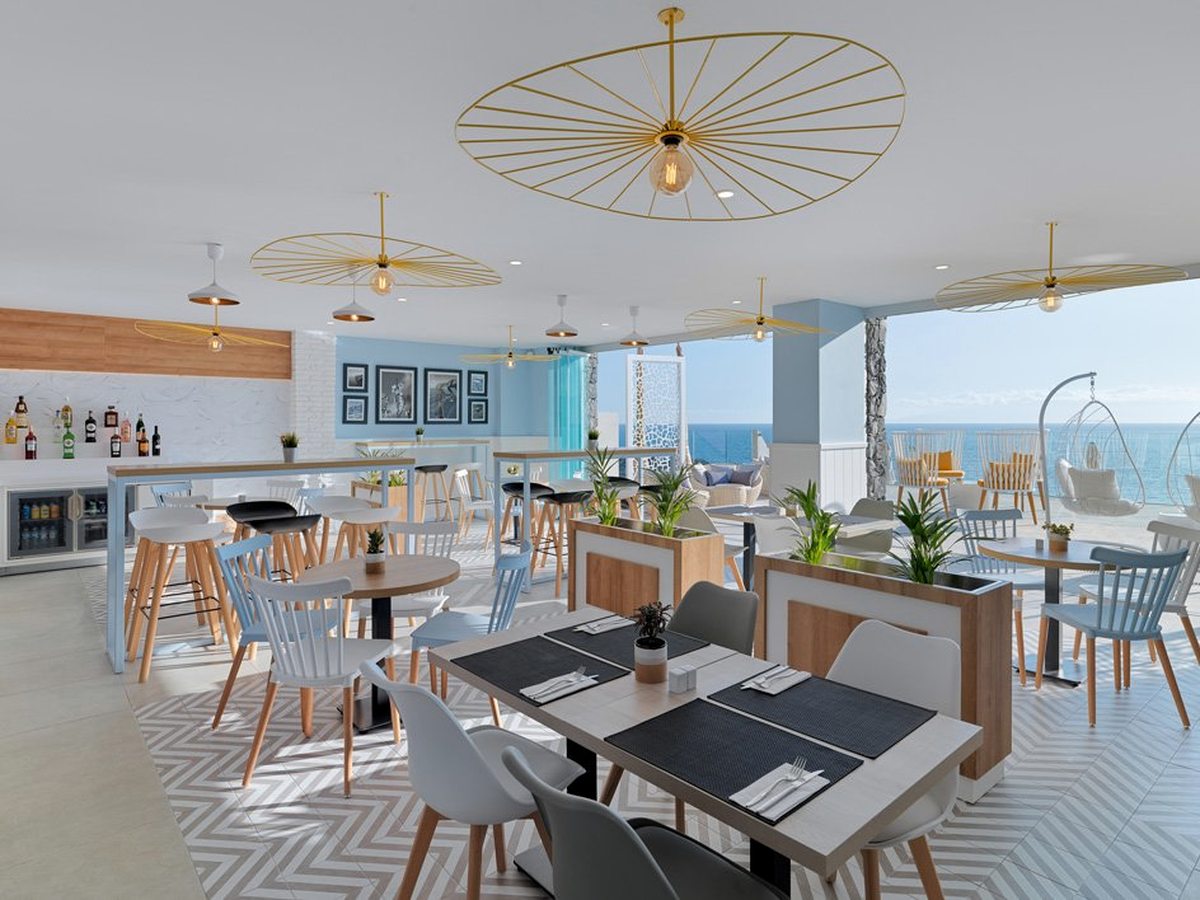 Indoor bar with a white counter and bottle shelves, pale wooden chairs at set tables under gold pendant lights, opening onto a terrace above the sea.