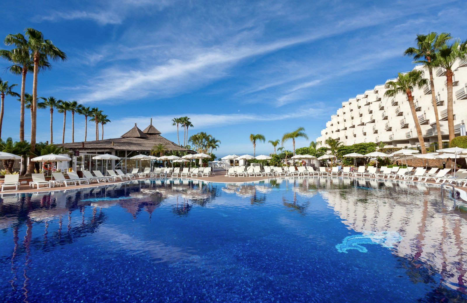 A large blue pool at Hotel Landmar Playa La Arena, edged with sun loungers and white parasols, with a thatched poolside bar among palms and the white terraced hotel building rising on the hillside behind.